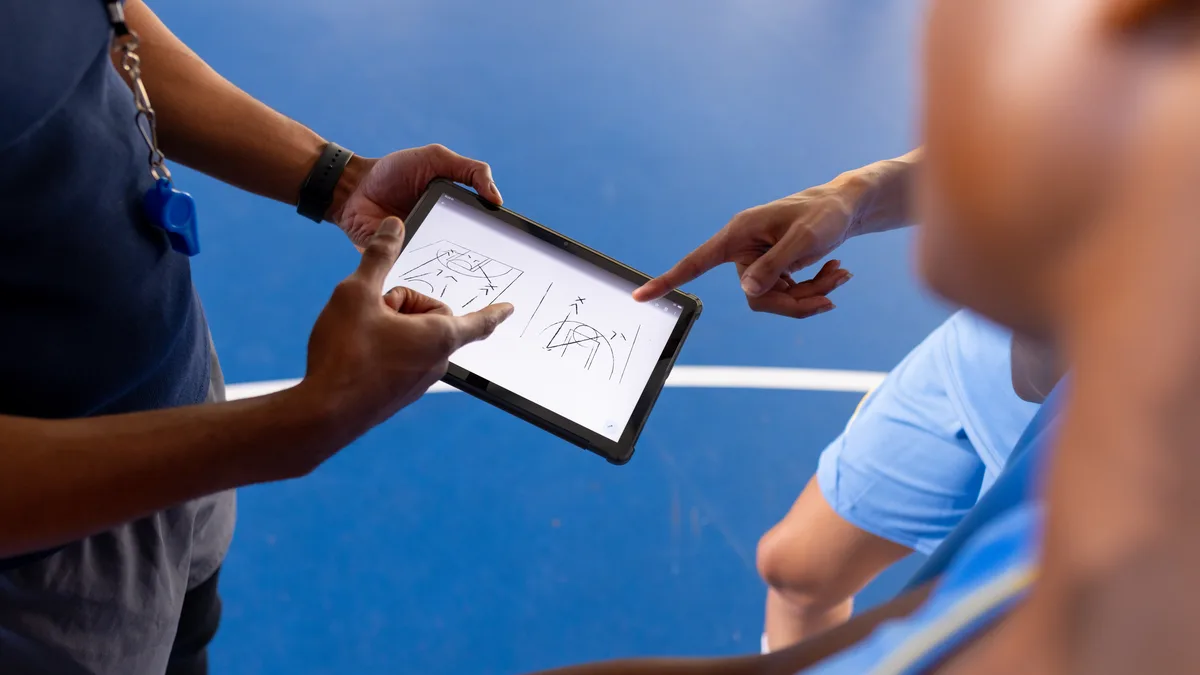 Un entraîneur montre un schéma de jeu sur une tablette, un joueur pointe l'écran du doigt, au bord d'un terrain bleu