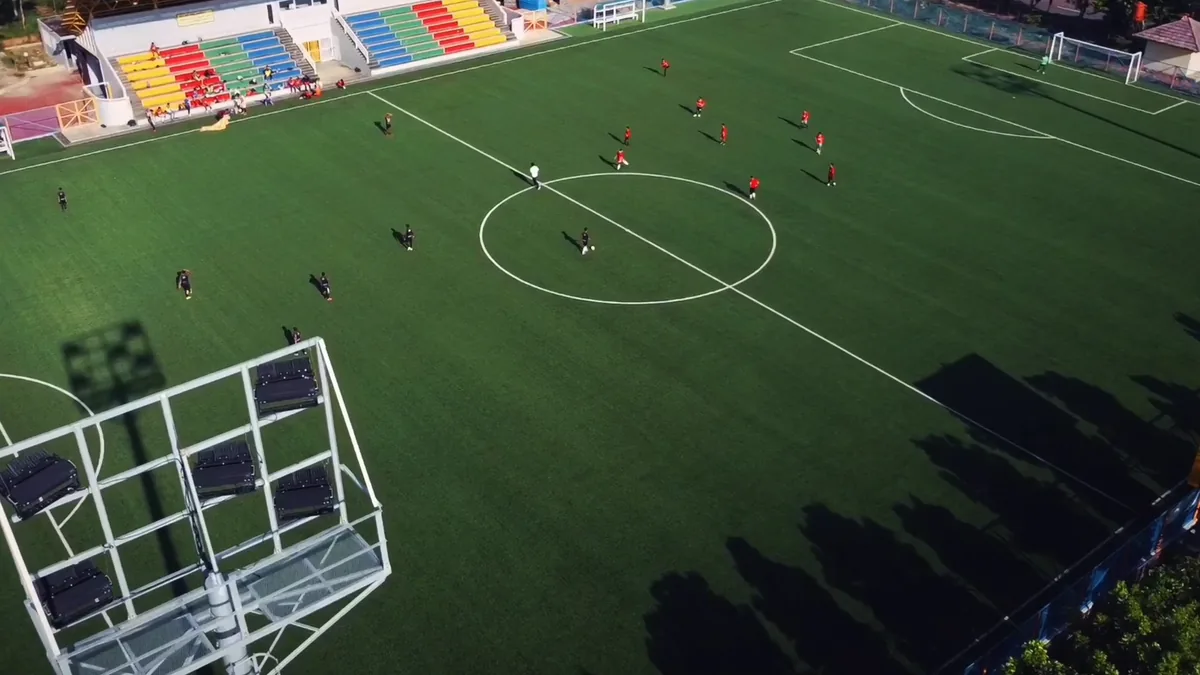 Match de football amateur vu du haut d'un mât d'éclairage, tout le terrain dans le cadre, tribune colorée au fond