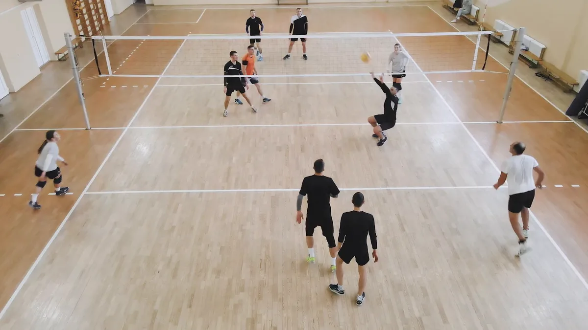 Match de volley amateur filmé depuis la galerie d'un gymnase, un passeur sous le ballon près du filet