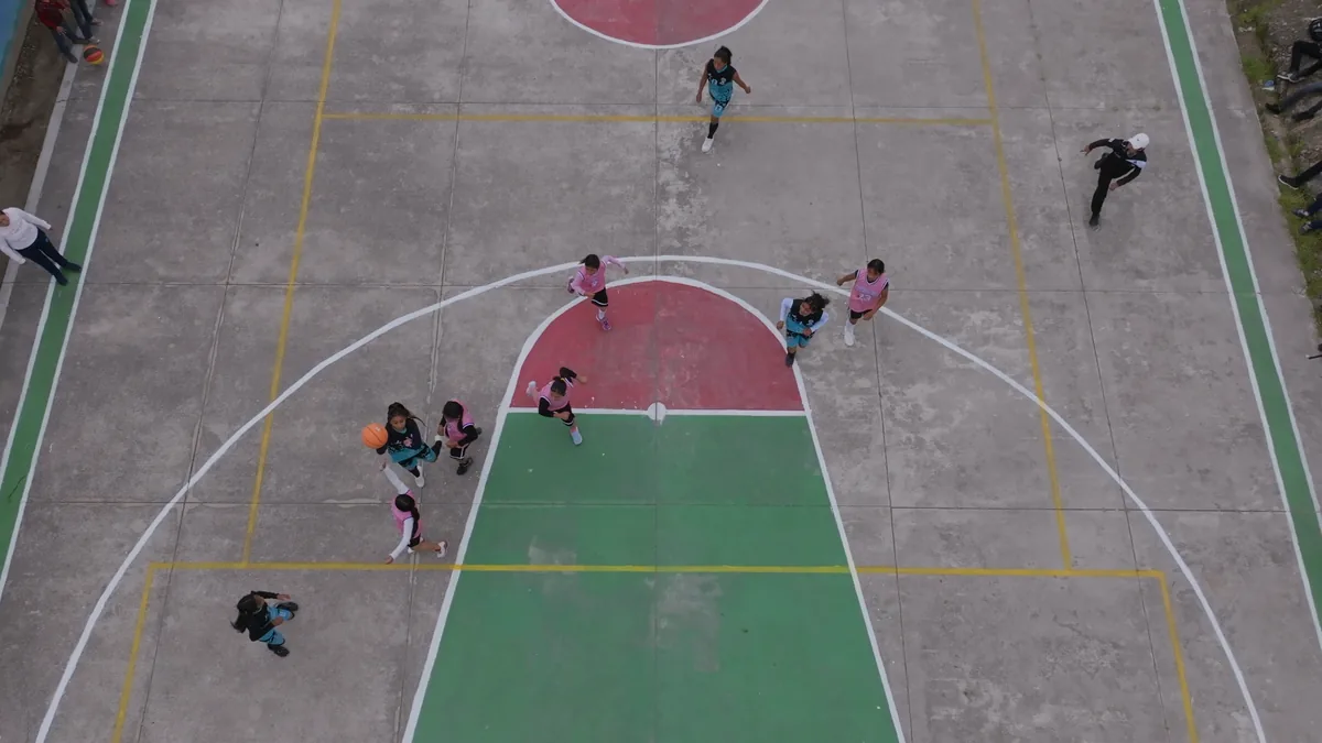 Match de basket de jeunes joueuses vu de haut sur un terrain extérieur, maillots roses et turquoise, ballon près de la raquette