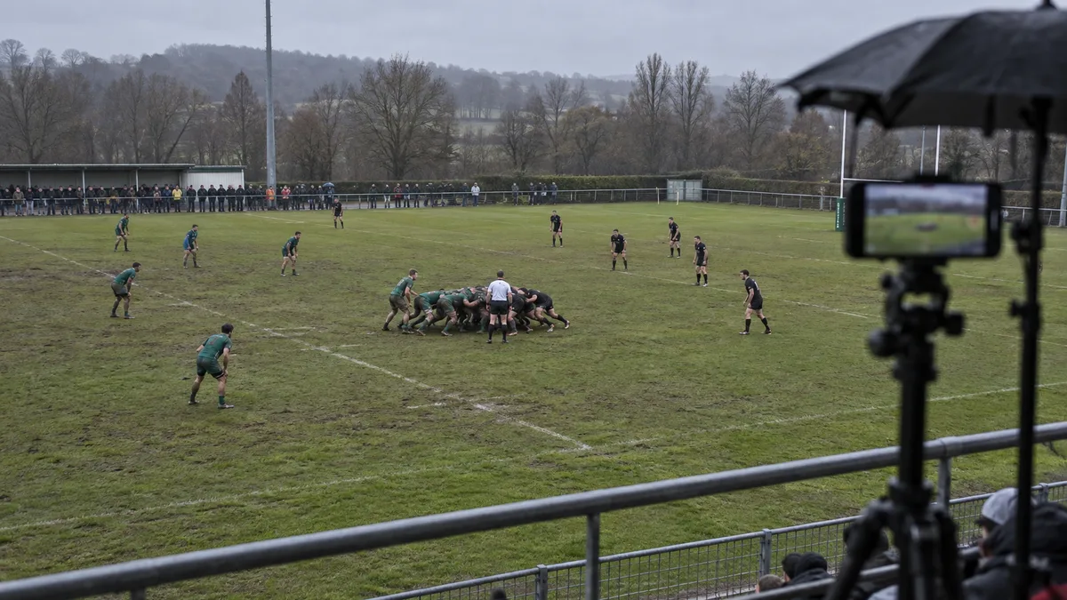 Téléphone sur trépied en haut d'une tribune de rugby amateur sous un ciel gris, mêlée en cours sur un terrain humide