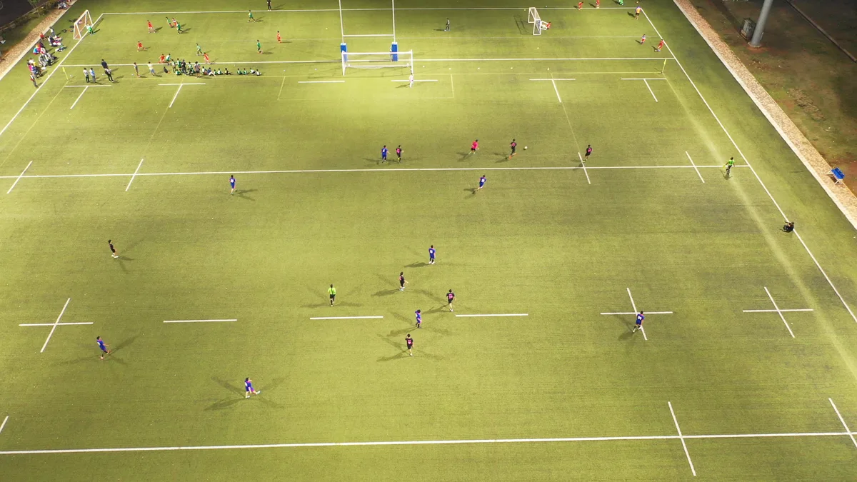 Match sur un terrain synthétique éclairé en fin de journée, vu d'en haut, joueurs en bleu et en noir étalés sur toute la largeur