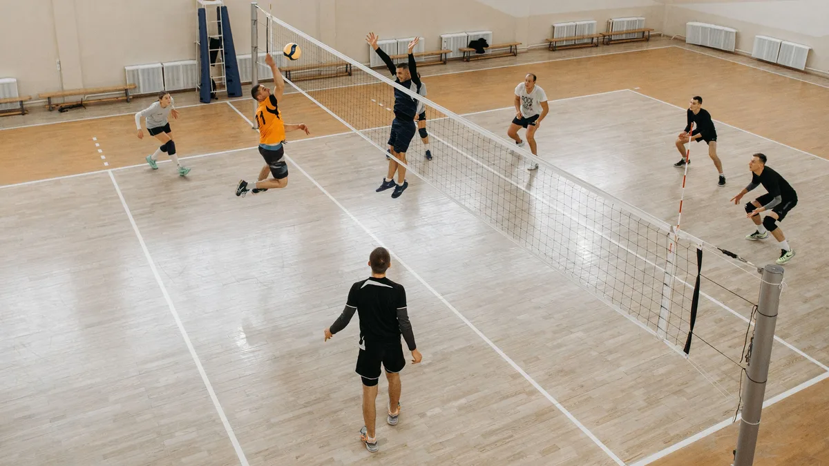 Match de volley amateur vu depuis la galerie d'un gymnase, un attaquant en orange frappe le ballon face à un contreur au filet