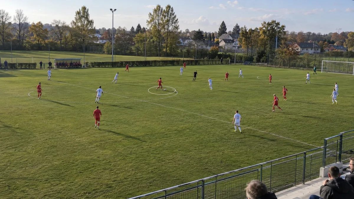 Plan large d'un match de football amateur filmé depuis le haut d'une tribune, un joueur en rouge ballon au pied dans le rond central